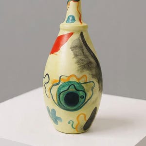 Peut inclure: Un vase en céramique peint à la main, avec un col étroit et un corps arrondi. Le vase présente des motifs abstraits rouges, noirs, bleus et oranges sur un fond crème. Le vase est exposé sur une surface blanche.