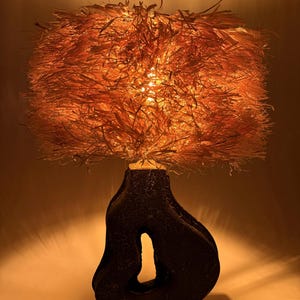 K&ouml;nnte beinhalten: Dekorative Tischlampe mit einem strukturierten, dunkelbraunen Sockel und einem gro&szlig;en, orangefarbenen, gefransten Lampenschirm. Die Lampe strahlt ein warmes, einladendes Licht aus. Der Sockel hat eine einzigartige, organische Form mit einer gro&szlig;en &Ouml;ffnung.