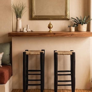 Puede incluir: Dos taburetes de bar de madera negra con asientos de paja tejida. Los taburetes tienen un diseño de escalera en las patas. Los taburetes están colocados frente a una pared de color neutro, con una estantería de madera y objetos decorativos en el fondo.