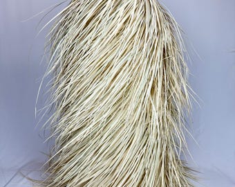 May include: A natural-colored, woven grass mask with a rounded top and long, flowing strands. The mask has a simple, organic design, with a small metal loop at the top for hanging. The overall aesthetic is rustic and earthy.