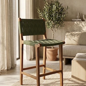 Op de afbeelding: Een hoge barkruk met een lichtbruin houten frame en een gevlochten zitting en rugleuning in een donker olijfgroene kleur. Het gevlochten materiaal vertoont een herhalend geometrisch patroon over zowel de zitting als de rugleuning.
