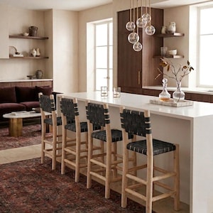 May include: Four bar stools with light-toned wooden frames and black woven leather seats and backrests. The stools feature a tall, sturdy design with horizontal footrests, positioned along a white kitchen island with a smooth, solid surface.