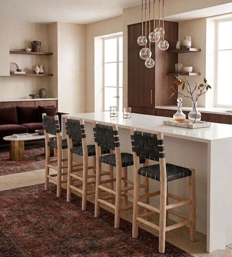 May include: Four bar stools with light-toned wooden frames and black woven leather seats and backrests. The stools feature a tall, sturdy design with horizontal footrests, positioned along a white kitchen island with a smooth, solid surface.