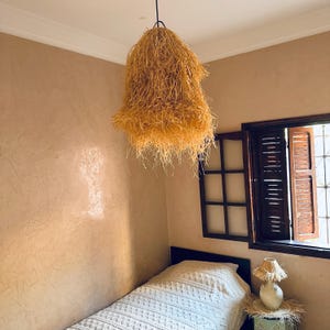 May include: A hanging, bell-shaped light fixture made of dried, straw-like material. The fixture is suspended from the ceiling in a room with tan walls and a window with wooden shutters. A bed with a white blanket is visible below.