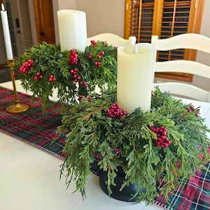 May include: Two white pillar candles sit atop green wreaths adorned with red berries. The wreaths are placed on a white table with a red and green plaid runner. A gold candlestick with a white candle is visible.