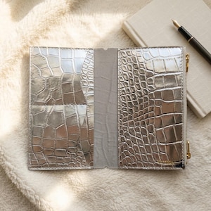 May include: A metallic silver wallet with a textured crocodile skin pattern, featuring an open interior with multiple card slots and a side zipper closure. The wallet is displayed flat, showcasing its reflective surface and structured design.