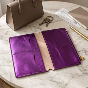 May include: A metallic purple leather wallet featuring an open interior with multiple card slots on the left side and a zippered compartment on the right side, finished with a small gold-toned heart-shaped zipper pull for secure closure.