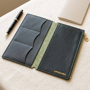 May include: A dark green leather notebook cover featuring three interior card slots, a zippered compartment with a heart-shaped pull, and the name ANURADHA embossed in gold. A black fountain pen with gold trim rests beside the open leather cover.