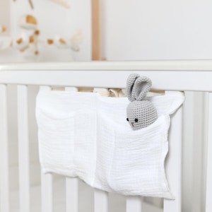 Puede incluir: Un organizador de cuna de tela blanca texturizada con tres bolsillos, sujeto a la barandilla de la cuna. Un pequeño juguete de conejito de ganchillo gris está en uno de los bolsillos. Un móvil con detalles amarillos y blancos es visible en el fondo.