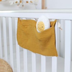 Può includere: Un organizer per culla giallo senape con due tasche è appeso a una culla bianca. Un uovo di peluche bianco e giallo con una faccia sorridente è in una tasca. Un mobile con figure di uccelli bianchi è sullo sfondo.