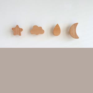 May include: Four wooden wall hooks in the shapes of a star, cloud, raindrop, and crescent moon. The hooks are light brown and are arranged in a row on a white background.