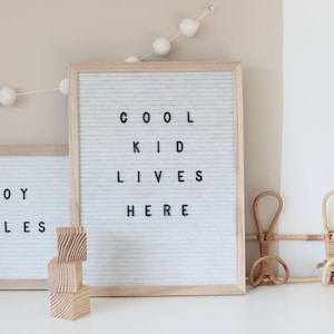 Pode incluir: Um quadro de letras de feltro emoldurado com o texto "COOL KID LIVES HERE" em letras pretas. O quadro está em uma moldura de madeira clara. Há três blocos de madeira empilhados em frente ao quadro. Uma guirlanda de pompons brancos pendurada acima.