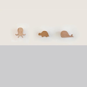 Puede incluir: Tres decoraciones de pared de madera con criaturas marinas: un pulpo, una tortuga y una ballena. Cada una está tallada en madera de color claro y tiene un diseño simple y minimalista. El pulpo tiene ocho tentáculos, la tortuga tiene un caparazón redondeado y la ballena tiene un cuerpo curvado.