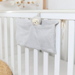May include: A grey baby comforter with a stuffed animal head attached, hanging on a white cot. The comforter has a soft, crinkled texture. A mobile with white and gold accents is visible in the background.