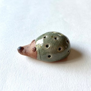 以下が含まれることがあります： 緑色の釉薬と茶色のアクセントが施された、小さな陶器のハリネズミの置物。ハリネズミは丸い体に穴が空いており、小さなとがった鼻をしています。