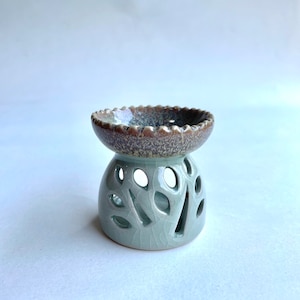 May include: A light green ceramic oil burner with a brown and white top. The burner has a decorative design of leaves cut out of the sides.