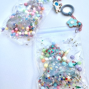 Op de afbeelding: Twee doorzichtige plastic zakken gevuld met kleurrijk glitter, kralen en sterren. De zakken bevatten een blauwe en witte wolkvormige harsbedel en een heldere harsbedel met glitter en sterren. Een zilveren ketting met een blauwe bloemenbedel en een astronautbedel is bevestigd aan de bovenkant van de zakken.