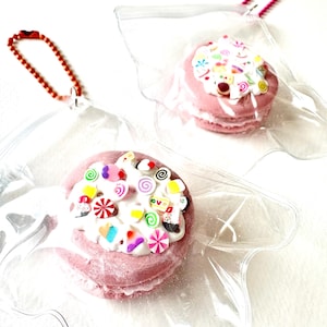 Op de afbeelding: Roze macaronvormige sleutelhangers met witte glazuur en kleurrijke snoepdecoraties. Elke sleutelhanger is verpakt in een doorzichtige plastic zak met een rode of roze kralenketting.