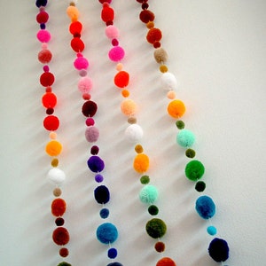 May include: Five strands of colorful pom-pom garland hanging on a white wall. The garlands are made of various colors, including red, orange, yellow, green, blue, purple, pink, and white.