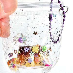 May include: A clear, flexible plastic pouch filled with black glitter and various colorful charms, including small flowers, a sun, and a green swirl. Inside the pouch sits a miniature brown and tan pudding dessert. A purple ball chain is attached.