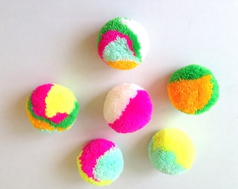 Può includere: Sei pompon colorati in varie tonalità di verde, rosa, giallo e bianco. I pompon sono fatti di filato e hanno una consistenza soffice.