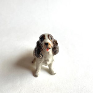 May include: A small ceramic dog figurine in a sitting position. The dog has a white body with brown and black markings on its head and ears. Its tongue is visible, and it has a glossy finish.