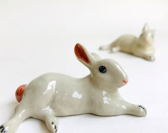 May include: Two ceramic rabbit figurines. The foreground rabbit is white with a pink nose, red-orange ears, and a small red-orange tail. The rabbit has blue eyes and black paw details. The second rabbit is in the background.