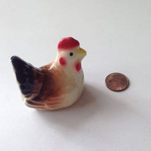 May include: A small ceramic chicken figurine with brown and black markings. The chicken has a red comb and a yellow beak.
