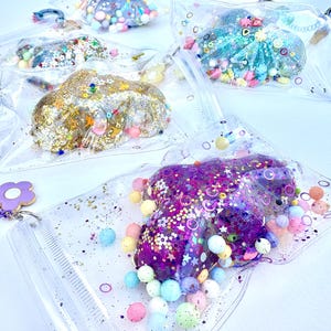 May include: Four clear plastic bags filled with colourful glitter, beads, and sequins. Each bag has a different colour theme: gold, purple, blue, and white.