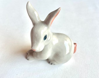 May include: A small, white ceramic rabbit figurine with blue eyes and pink accents. The rabbit is sitting on its hind legs with its front paws resting on the ground.