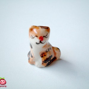 May include: A small ceramic figurine of a cat with brown, white, and black markings. The cat has a closed-eye smile and a red nose.