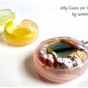 Puede incluir: Una funda de gelatina rosa translúcida para un dispositivo de mascota digital con una cadena de bolas de metal, acompañada de dos recipientes circulares de gelatina más pequeños en amarillo y verde. El texto Jelly Cases for Originals by iammie es visible.