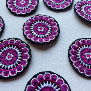 Könnte beinhalten: Ein Set mit zehn gestickten Bügelbildern in Lila, Weiß und Schwarz. Jedes Bügelbild zeigt ein kreisförmiges Mandala-Design mit einem rosafarbenen Zentrum.