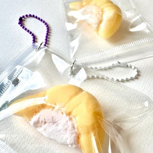 May include: Close-up of two clear plastic pouches, each containing a yellow and white shrimp-shaped item. One pouch has a purple beaded chain, the other a silver beaded chain. The background is a white surface.
