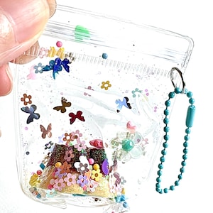 May include: A clear, flexible plastic pouch filled with colorful confetti, including small flowers, butterflies, and glitter. The pouch features a zipper closure at the top and a turquoise ball chain attached to a metal ring on the side for hanging.