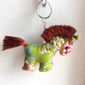 Pode incluir: Um chaveiro de cavalo de tecido verde com um padrão floral marrom e uma cauda de borla marrom. O cavalo tem uma crina marrom e um chaveiro prateado.