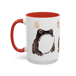 May include: White ceramic coffee mug with a red rim. The mug features a black and white illustration of a frog with a Japanese character on the side.