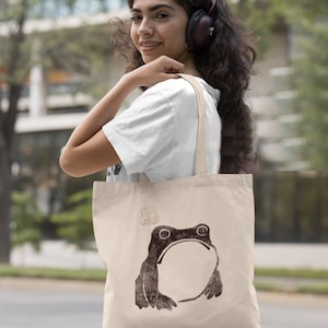 May include: A beige tote bag with a brown frog illustration and a small brown stamp with Japanese characters. The bag has a long strap and is being held by a person.