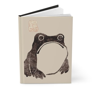 May include: A beige journal with a black spine and a black and brown frog illustration on the front cover. The frog is facing forward with its mouth closed. The journal has a red stamp with Japanese characters on the front cover.