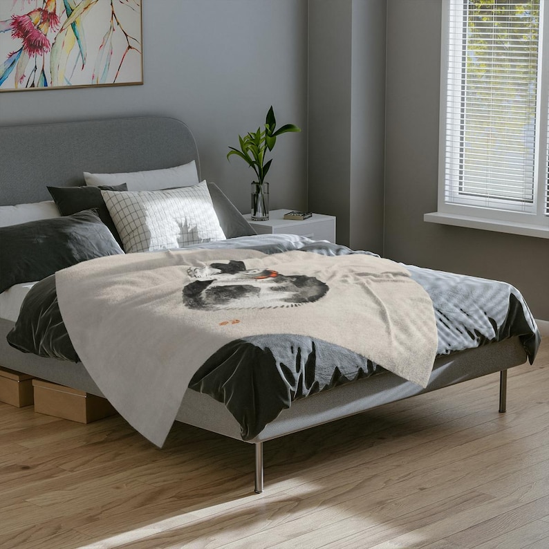 May include: A beige throw blanket with a black and white illustration of a cat curled up on a bed. The blanket is on a gray bed with black and white pillows.