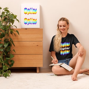 May include: A black t-shirt and a framed print with the word "Optics" in a rainbow gradient. The t-shirt is worn by a person sitting on a white rug. A wooden cabinet and a green plant are also in the image.