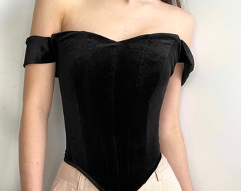 May include: A black velvet corset top featuring an off-the-shoulder neckline with short sleeves and a pointed hemline. The bodice is constructed with vertical seam detailing that creates a structured shape, paired with light beige trousers.