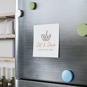 May include: A white square magnet with the text "Jot & Java" and a coffee cup design in brown, attached to a stainless steel refrigerator door. The phrase "IDEAS START HERE" is below the logo. Several round magnets in green, white, and blue are also on the door.