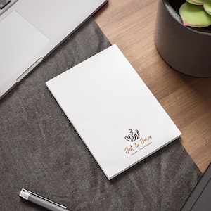 Könnte beinhalten: Ein weißer Notizblock mit dem Text "Jot & Java IDEAS START HERE" in Braun. Der Notizblock liegt auf einem Holztisch neben einem Laptop, einem Stift und einer Topfpflanze.