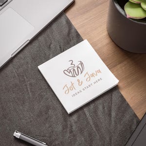 Puede incluir: Un bloc de notas adhesivas blancas con el texto "Jot & Java IDEAS START HERE" en marr&oacute;n y beige, y un logotipo de taza de caf&eacute;. Las notas est&aacute;n sobre una superficie gris oscura, junto a un port&aacute;til y un bol&iacute;grafo.