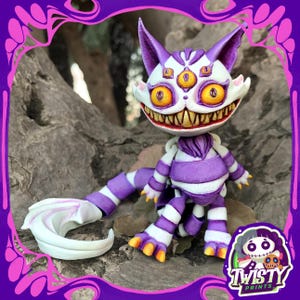 Puede incluir: Una caprichosa figura del Gato de Cheshire, morada y blanca, con tres ojos y una gran sonrisa. El gato tiene patas y cola a rayas, con garras y detalles en naranja. De "Twisty Prints".
