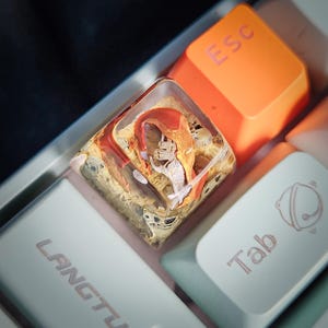 May include: A clear resin keycap with a red and orange abstract design. The keycap is on a white keyboard with the keys "Esc" and "Tab" visible.