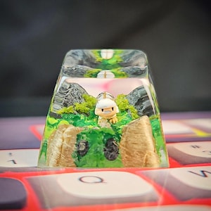 May include: A miniature figurine of a character with a white helmet and brown body is encased in a clear resin keycap. The keycap is filled with green moss, brown rocks, and a green and brown painted landscape.