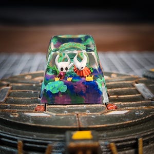 Może przedstawiać: Przezroczysty klawisz z żywicy z kolorowym, abstrakcyjnym wzorem. Wewnątrz klawisza znajdują się dwie małe, białe figurki postaci z gry Hollow Knight.