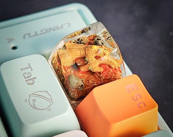 May include: A clear resin keycap with a gold-colored figurine of a hand with colorful stones embedded in it. The keycap is on a light blue keyboard with the keys "Tab" and "Esc" visible.